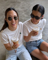 Two people sitting on a concrete floor, wearing sunglasses and white t-shirts, holding coffee cups.